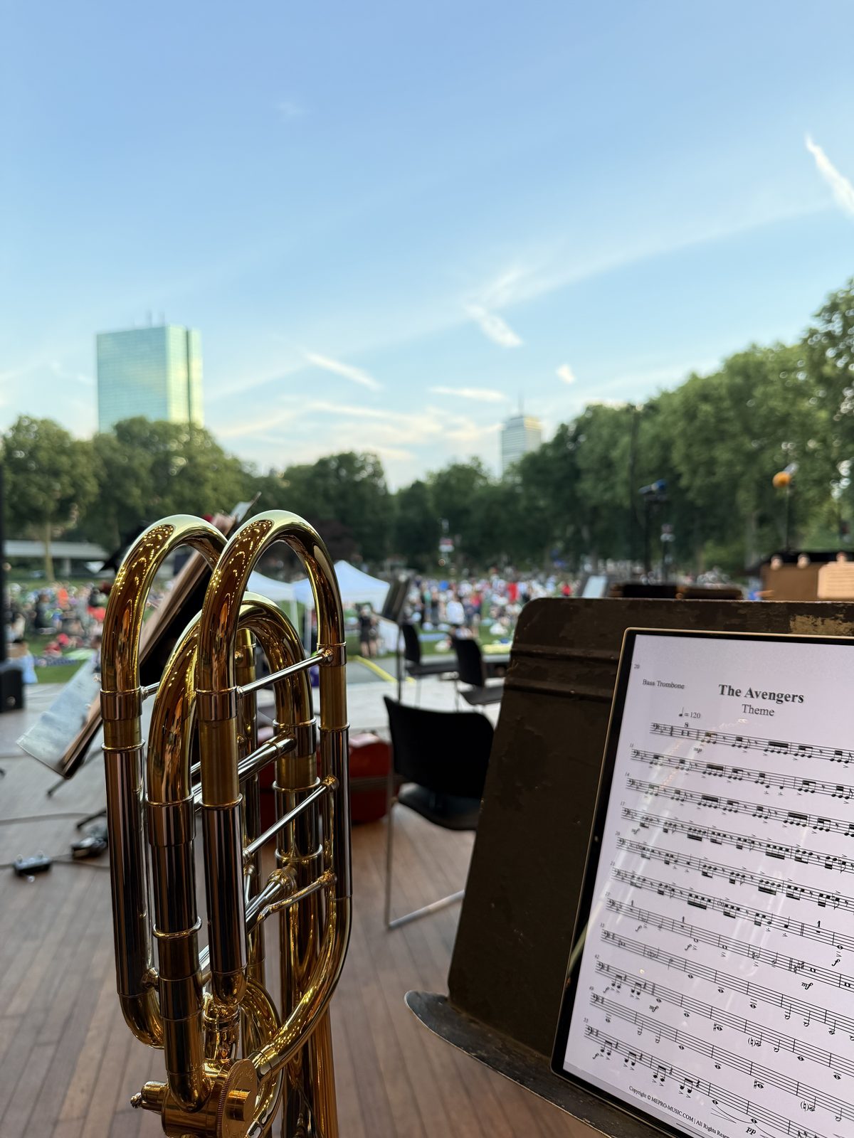 An HP Envy x360 showing a bass trombone part, on a music stand at an outdoor concert, with a trombone in the foreground and the audience on the grass beyond.